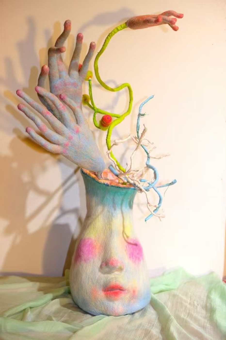 Past vessel-like soft sculpture with face, pale textile hands, and branching colored stems.