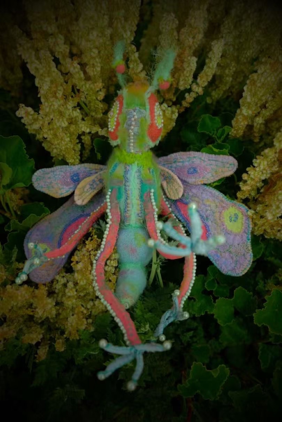 Past insect-like textile creature with soft wings photographed among dark green flowers.