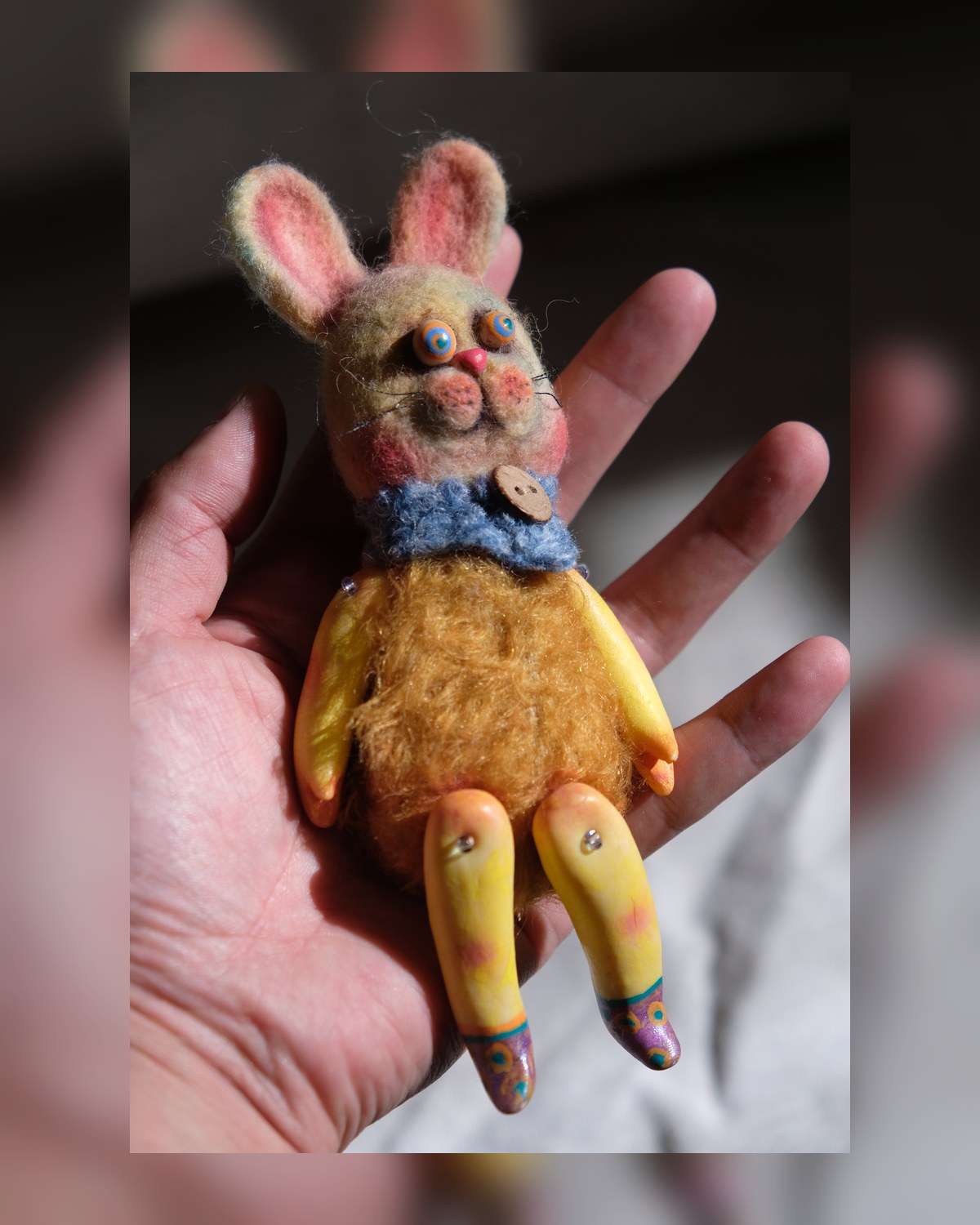 Front view of Tick Tock resting in a hand, showing its rabbit face, wool body, blue collar, and jointed legs.